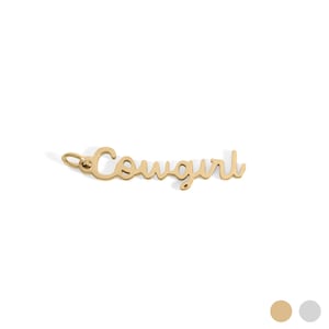 Puede incluir: Un colgante dorado con la palabra "Cowgirl" en cursiva. El colgante tiene un pequeño lazo en la parte superior para sujetarlo a una cadena. El fondo es blanco.