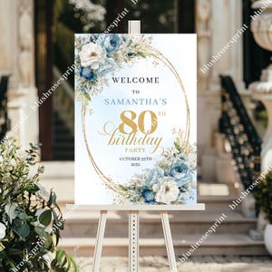 May include: A sign for Samantha's 80th birthday party, displayed on an easel. It features a gold oval frame with blue and white floral decorations. The text reads "Welcome to Samantha's 80th Birthday Party, October 25th, 2031."