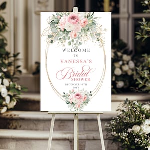 May include: A white sign on an easel with the text "WELCOME TO VANESSA'S Bridal SHOWER DECEMBER 16TH 20YY". The sign is decorated with pink and white flowers, green leaves, and gold accents. The sign is on a white easel.