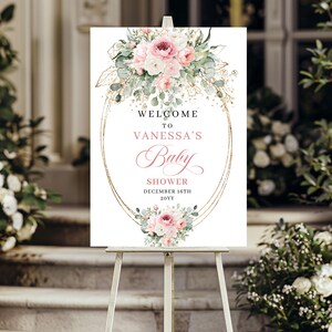 May include: A white sign on an easel with floral arrangements and gold accents. The sign reads "WELCOME TO VANESSA'S Baby SHOWER DECEMBER 16TH 20YY". The floral arrangements include pink and white roses and greenery.