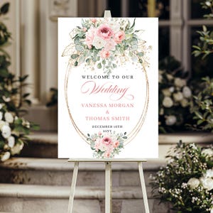 May include: A wedding welcome sign on an easel. The sign features a floral arrangement with pink and white flowers, greenery, and gold accents. The text reads "Welcome to Our Wedding" and includes the names Vanessa Morgan & Thomas Smith, and the date December 16th.