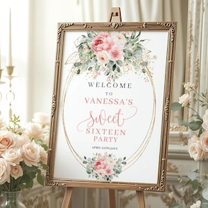 May include: A framed sign with a floral design and the text "Welcome to Vanessa's Sweet Sixteen Party." The sign is in a gold frame and is displayed on a wooden easel. The floral arrangement includes pink and white flowers.