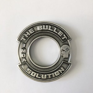 Puede incluir: Llavero de metal plateado con un anillo en el centro. El llavero tiene las palabras "The Bullet Solution" y "1 MOA" alrededor del anillo. Un cráneo con una mira sobre un ojo está en el lado derecho del anillo.
