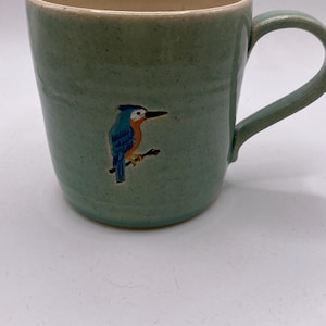 May include: A light green ceramic mug with a speckled finish and a handle on the right side. The exterior features a small, detailed illustration of a blue and orange kingfisher bird perched on a thin brown branch, centred on the mug body.