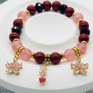 Könnte beinhalten: Ein Armband mit rosa und weinroten Perlen, durchsetzt mit schwarzen facettierten Perlen. Goldfarbene Charms umfassen zwei blütenförmige Anhänger und einen Charm mit herzförmigen Edelsteinen. Das Armband wird auf einer weißen Oberfläche präsentiert.