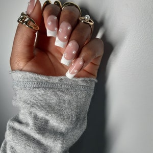 May include: A hand displaying long, square acrylic nails with a white French tip design and small, white pearls. The fingers are adorned with gold rings, including a chain-link style and a ring with a pearl.