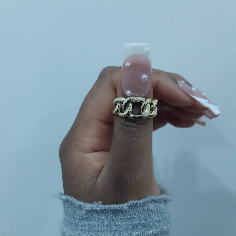 May include: A gold-coloured chain link ring. The ring features a chunky design with interlocking links. The ring is worn on a finger. The nails are painted with a light pink and white design.