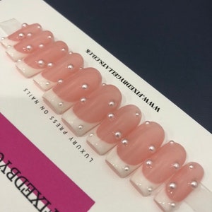 May include: A set of press-on nails featuring a French manicure design. The nails are pale pink with white tips and adorned with small pearl accents. The text "FIXEDBYGELLATS.CO.UK" is visible.