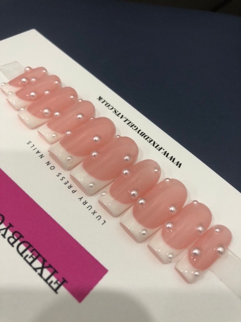 May include: A set of press-on nails featuring a French manicure design. The nails are pale pink with white tips and adorned with small pearl accents. The text "FIXEDBYGELLATS.CO.UK" is visible.