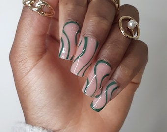 May include: A hand displaying square-shaped, pale pink nails with a green wavy pattern. Several gold rings adorn the fingers, including a chain link design and a ring with a pearl.