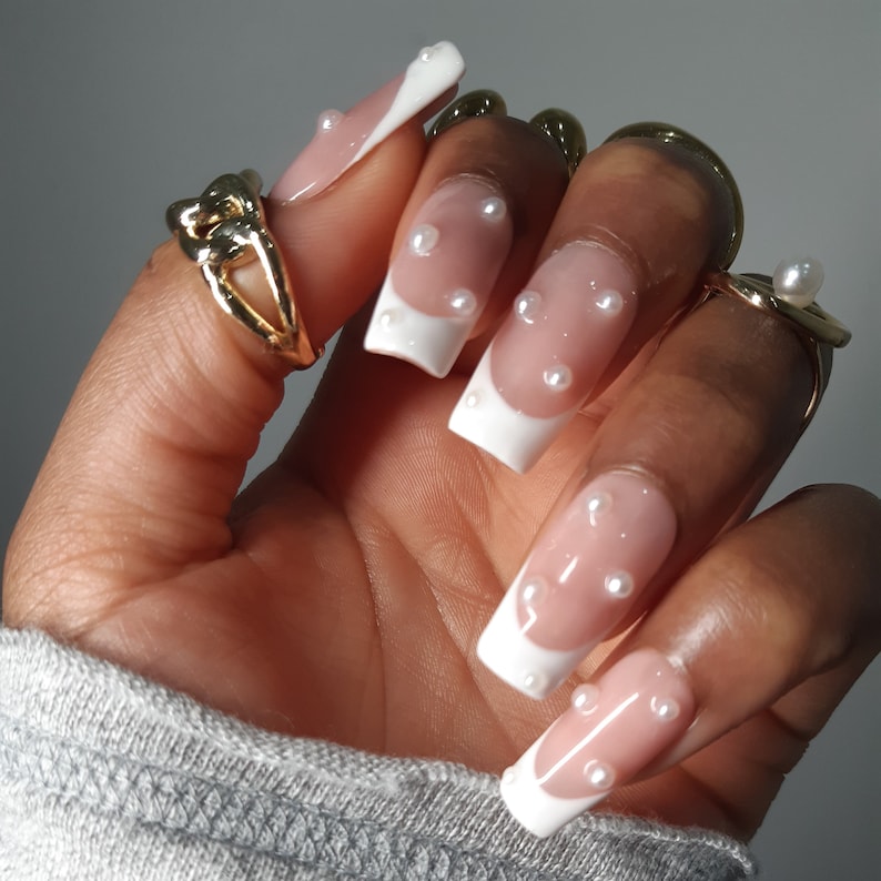 May include: A hand displaying long, square artificial nails. The nails have a white French tip design with small, white pearl embellishments. Gold rings are worn on the fingers.