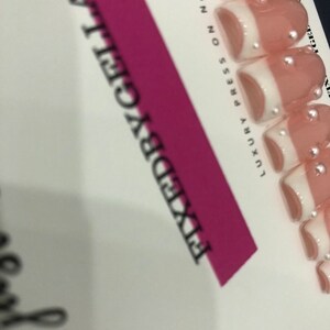 May include: A set of press-on nails with a French tip design. The nails are a light pink with white tips and small, silver embellishments. The packaging includes the text "FIXEDBYGELATTS" and "LUXURY PRESS ON NAILS".
