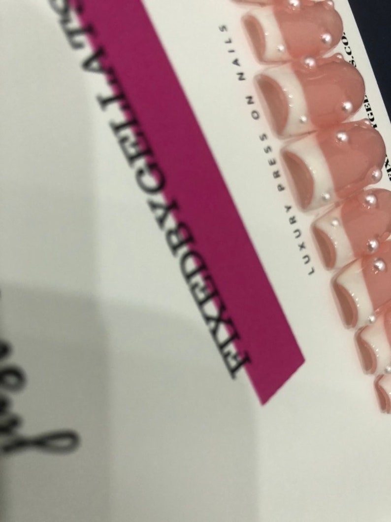 May include: A set of press-on nails with a French tip design. The nails are a light pink with white tips and small, silver embellishments. The packaging includes the text "FIXEDBYGELATTS" and "LUXURY PRESS ON NAILS".