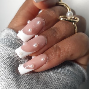 May include: Close-up of a hand with long, square nails. The nails have a French manicure design with white tips and a nude base, decorated with small, round pearls. A gold ring with a pearl accent is also visible.