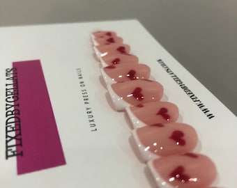 May include: A set of press-on nails with a white base and a pink, heart-shaped design. The nails are arranged on a white card with the text "FIXEDBYCELLATS" and "LUXURY PRESS ON NAILS".