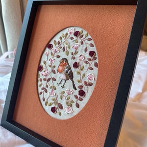 Könnte beinhalten: Gerahmte Stickerei mit einem Rotkehlchen, umgeben von rosa und burgunderfarbenen Blumen und grünen Blättern. Das Kunstwerk ist vor einem strukturierten, orangefarbenen Hintergrund platziert und in einem schwarzen Rahmen gefasst. Die Stickerei ist detailliert und farbenfroh.