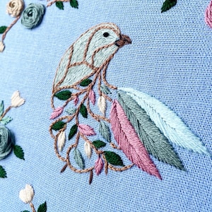 Könnte beinhalten: Gesticktes Kunstwerk mit einem Vogeldesign, das einen hellblauen Körper und eine braune Umrandung aufweist. Der Vogel ist mit rosa, grünen und weißen Blättern verziert. Der Hintergrund ist hellblauer Stoff, mit gestickten Blumen in den Ecken.