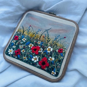 Può includere: Un telaio da ricamo quadrato contenente un pannello in tessuto con una scena paesaggistica cucita a mano. Il design presenta vivaci fiori rossi e bianchi su un prato verde scuro, con uno sfondo di cielo dipinto in tenui toni blu e arancioni.