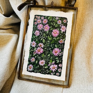 Könnte beinhalten: Ein gerahmtes Blumen-Aquarellgemälde. Das Kunstwerk zeigt rosa Blüten mit gelben Zentren und grünen Blättern vor einem dunklen Hintergrund. Der Rahmen ist goldfarben mit einem dunkelbraunen Samtband zum Aufhängen.