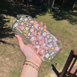May include: A clear protective case for a handheld gaming console featuring a glittery finish and various small, colorful, raised charms including stars, hearts, and sweets. The text "maryssi" is printed in white on the left side of the case.