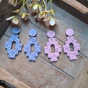 Op de afbeelding: Vier handgemaakte geometrische oorbellen in lavendel- en roze tinten. Elke oorbel heeft een textuurpatroon en een cirkelvormige bovenkant. De oorbellen worden gepresenteerd op een houten dienblad met bloemaccenten.