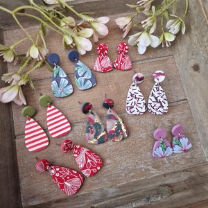 Puede incluir: Una colección de pendientes de arcilla polimérica hechos a mano en varios diseños y colores. Los pendientes incluyen patrones florales, a rayas y abstractos. La paleta de colores incluye rojo, blanco, verde, azul y rosa. Los pendientes se exhiben en una bandeja de madera con detalles florales.
