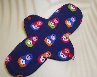 Puede incluir: Una compresa de tela reutilizable azul oscuro con un estampado colorido de muñecas rusas. La compresa tiene un cierre de presión y una pequeña etiqueta con la letra "H" en ella.
