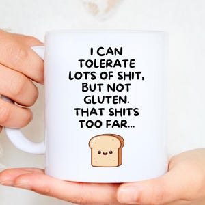 Puede incluir: Taza de cerámica blanca con el texto "I CAN TOLERATE LOTS OF SHIT, BUT NOT GLUTEN. THAT SHITS TOO FAR..." y una rebanada de pan de dibujos animados. La taza está sostenida por una persona.