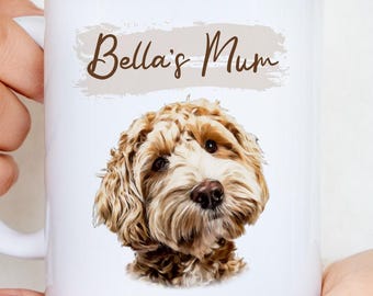 May include: A white ceramic mug featuring a brown and cream-colored doodle dog illustration centered on the front. Above the dog, the text Bella's Mum is printed in a brown script font over a light beige, textured brushstroke background.