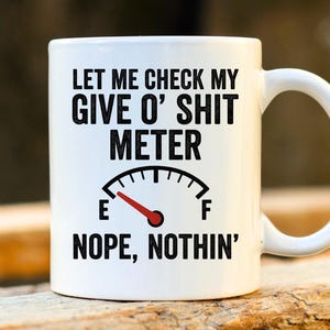 Könnte beinhalten: Weiße Keramik-Tasse mit schwarzem Henkel. Die Tasse zeigt den Text "LET ME CHECK MY GIVE O' SHIT METER" über einer Messanzeige. Darunter steht "NOPE, NOTHIN'".