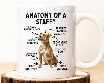 May include: A white ceramic mug with a handle, featuring the text "ANATOMY OF A STAFFY" and illustrations of a dog and its various functions, including "Snack Surveillance" and "Stink Dispenser". The mug sits on a wooden coaster.