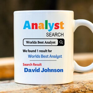 May include: White ceramic mug featuring the word "Analyst" in colourful letters. Below, the text reads "SEARCH" and "Worlds Best Analyst" within a search bar. The mug also displays "We found 1 result for Worlds Best Analyst" and "Search Result David Johnson."