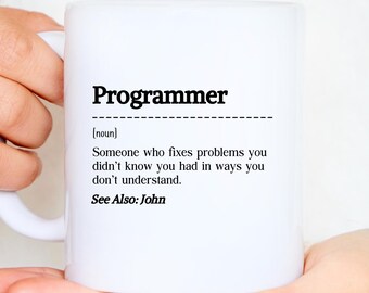 Peut inclure: Un mug en céramique blanc avec le mot "Programmer" en gras noir. En dessous, une définition : "Quelqu'un qui résout les problèmes que vous ne saviez pas avoir, de manières que vous ne comprenez pas." Le texte "See Also: John" est également présent.