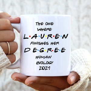 Puede incluir: Taza de cerámica blanca con texto negro: "The One Where Lauren Finishes Her Degree Human Biology 2021". El texto está escrito en una fuente similar a la de la serie de televisión Friends. La taza la sostiene una persona que lleva un jersey blanco.