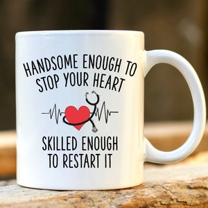 Può includere: Tazza in ceramica bianca con un designo nero e rosso. La tazza recita "Handsome enough to stop your heart" e "Skilled enough to restart it". Un cuore rosso con uno stetoscopio si trova al centro della tazza.