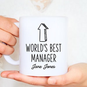 Może przedstawiać: Biały kubek ceramiczny z czarnym tekstem, który brzmi "World's Best Manager" i "Jane Jones". Nad tekstem narysowana jest strzałka skierowana w górę.