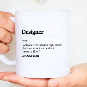 Op de afbeelding: Witte keramische mok met het woord "Designer" in zwart en een humoristische definitie van het beroep. De definitie luidt: "Iemand die acht uur besteedt aan het kiezen van een lettertype en het 'creatieve flow' noemt."