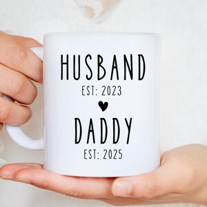 Puede incluir: Taza de cerámica blanca con texto negro que dice "Husband Est: 2023" y "Daddy Est: 2025" con un pequeño símbolo de corazón negro entre las dos líneas de texto.