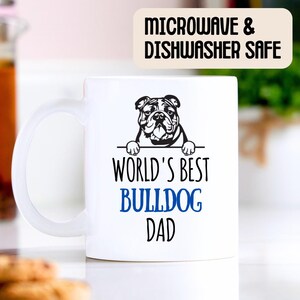 May include: White ceramic mug with a black and white illustration of a bulldog peeking over the top. The text "World's Best Bulldog Dad" is printed on the mug in black and blue. The mug is microwave and dishwasher safe.