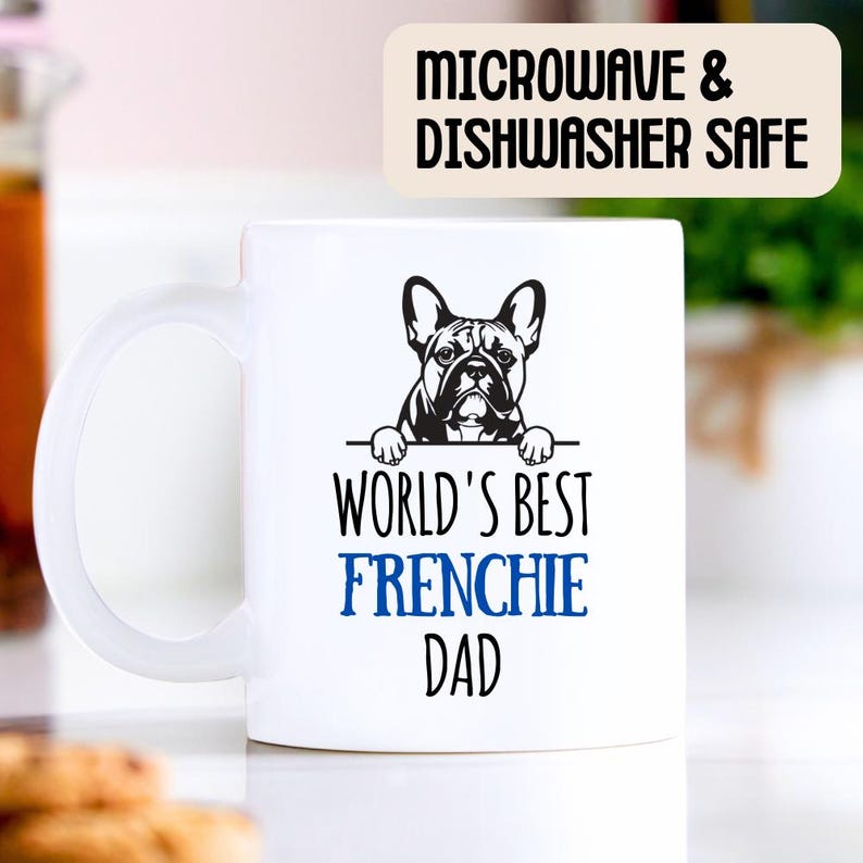 Op de afbeelding: Een witte keramische mok met een zwart-witte illustratie van een Franse Bulldog die over de rand kijkt. De tekst "World's Best Frenchie Dad" is in zwart en blauw op de mok gedrukt. De mok is geschikt voor de magnetron en vaatwasser.