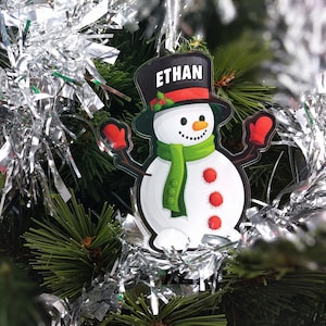 May include: A snowman Christmas bauble with a black top hat that says "ETHAN" on it. The snowman is wearing a green scarf and red mittens. The bauble is hanging on a Christmas tree with silver tinsel.