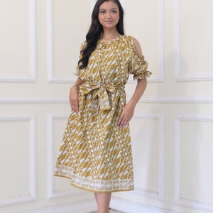 Puede incluir: Un vestido amarillo mostaza con un patrón blanco. El vestido tiene un diseño de hombros descubiertos y un lazo en la cintura.