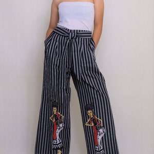 Könnte beinhalten: Schwarze und weiße, gestreifte, weite Hose mit einem schwarzen Gürtel und zwei gestickten Figuren auf den Beinen. Die Figuren tragen traditionelle indonesische Kleidung.