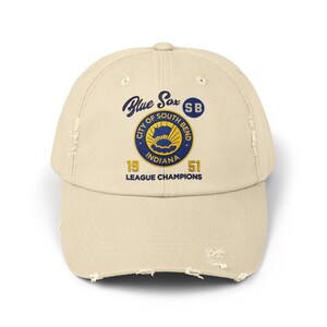 Op de afbeelding: Een beige, verweerde baseballpet met een blauw en goud geborduurd logo met de tekst "Blue Sox" en "City of South Bend Indiana 19 51 League Champions" met een blauwe cirkel met de tekst "SB" boven het logo.