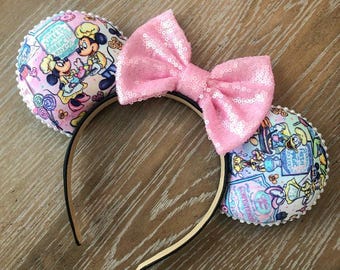 May include: A headband featuring two circular ears with a light pink, blue, and white pattern depicting cartoon characters in chef hats and sweets. A large, central, light pink sequined bow sits between the ears. The ears are trimmed with small white beads.