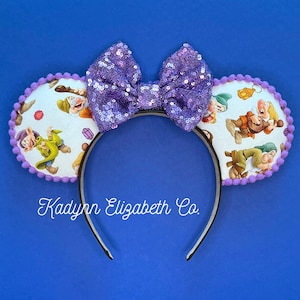 May include: Purple sequin bow headband with white fabric ears featuring the seven dwarfs from Snow White. The headband has a black band and the ears have a purple trim.