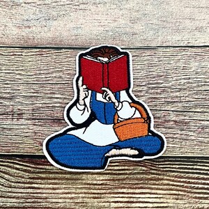 May include: Embroidered patch featuring a seated figure reading a red book. The figure wears a blue dress, white blouse, and has a brown basket. The patch has a white border and is likely for clothing or accessories.