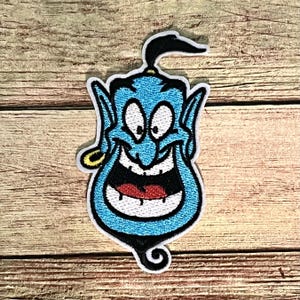 May include: A blue embroidered patch featuring a smiling genie with a black ponytail and a gold earring. The genie has a wide, open mouth with white teeth and a red tongue. The patch has a white border.