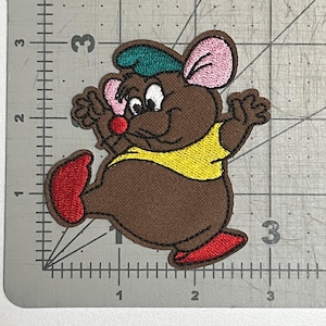 May include: A brown embroidered patch of a cartoon mouse wearing a green hat and a yellow shirt. The mouse is smiling and has pink ears.