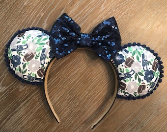 May include: A pair of blue and white mouse ears with a blue sequined bow. The ears are decorated with a floral pattern and footballs.
